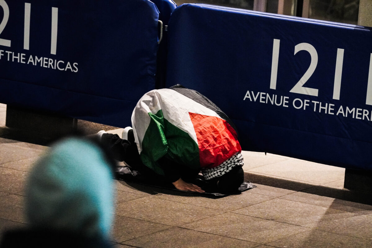 A protester wearing the palestinian flag as a cape and a keffiyeh tied around his neck touches his head to the ground in prayer 