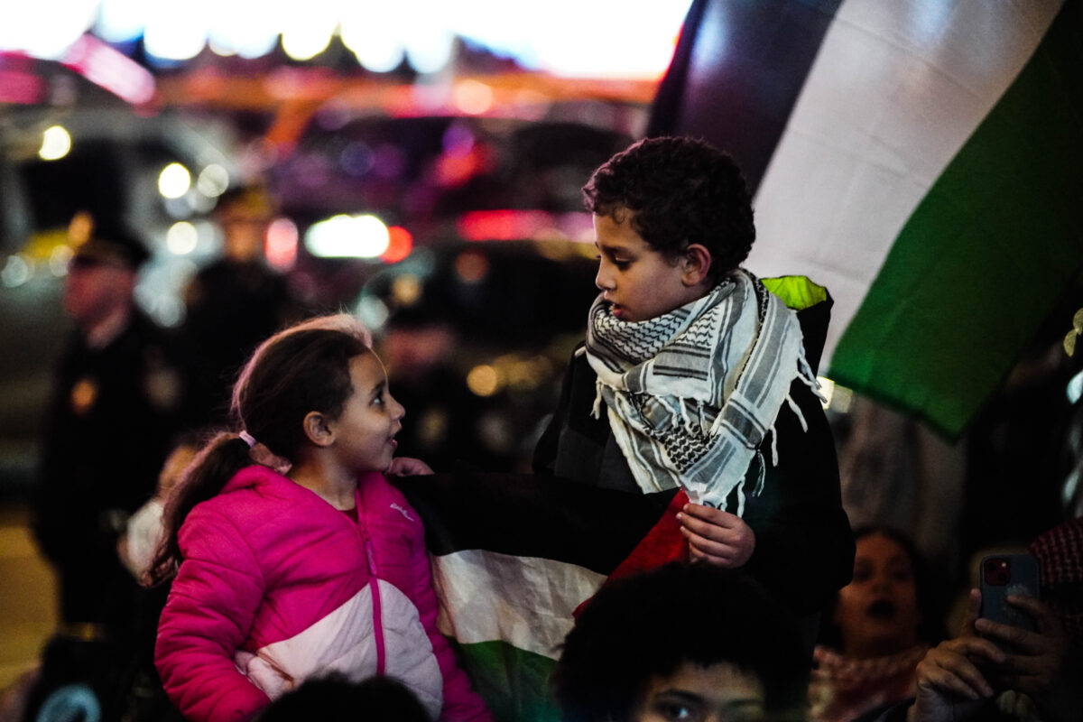 A young girl wearing pink (left) looks up at a young boy wearing a keffiyeh and holding the Palestinian flag (right)
