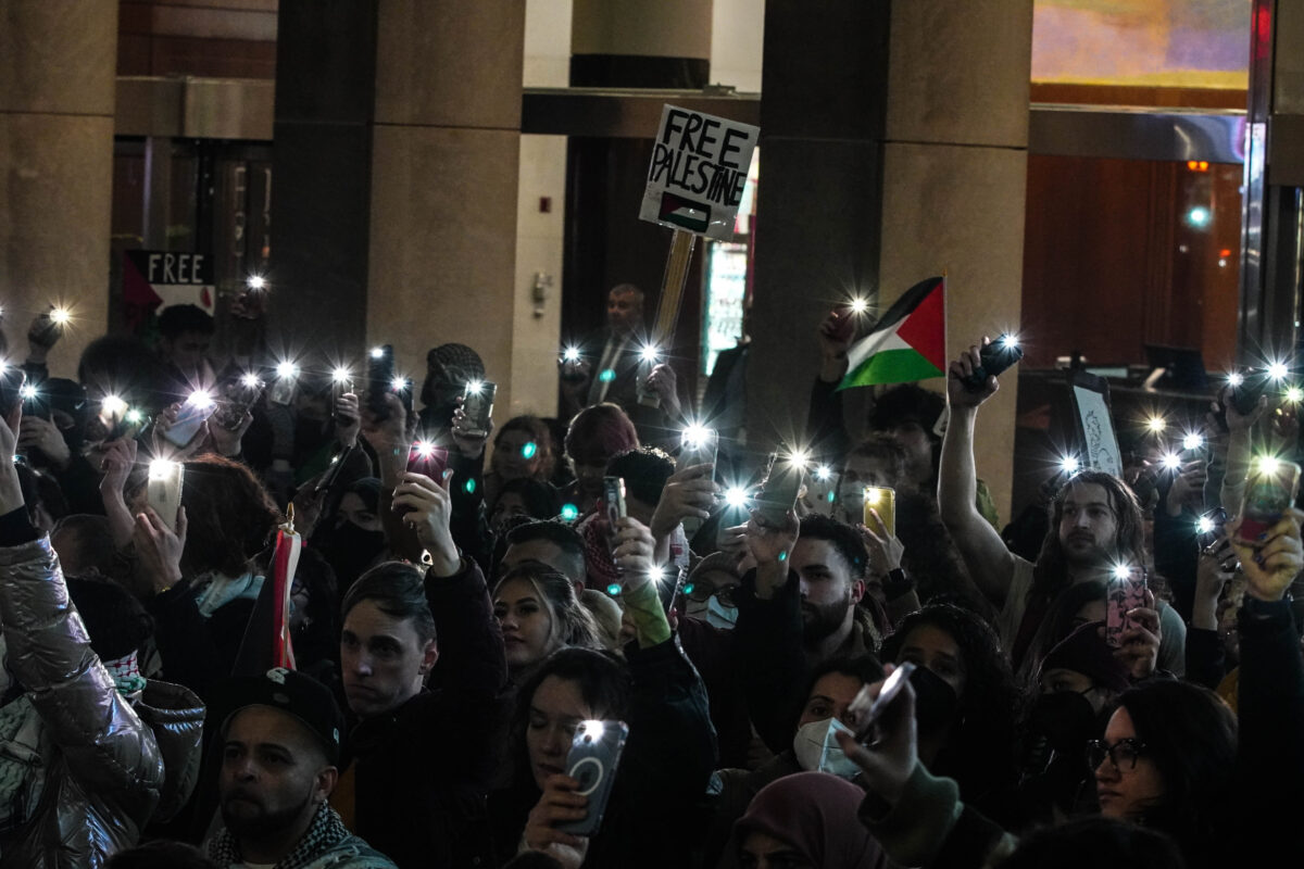 Group of protesters hold up their phone camera lights, signs, and flag of Palestine 