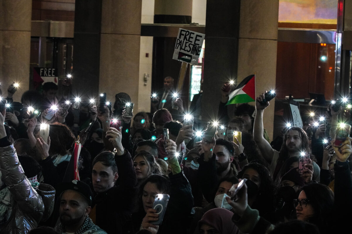 Group of protesters hold up their phone camera lights, signs, and flag of Palestine