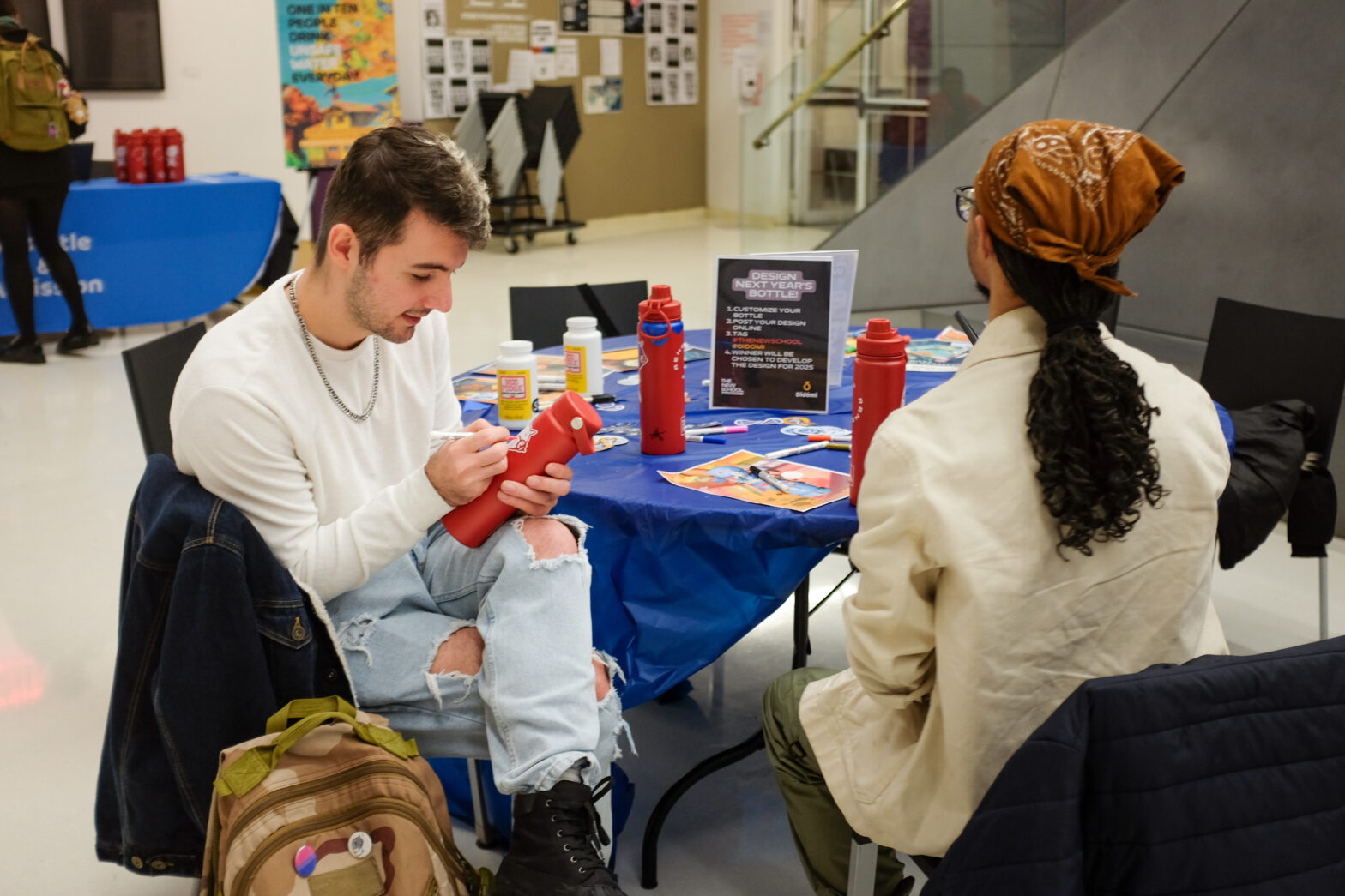 Two students sit at the water bottle customization table in the University Center’s event cafe, surrounded by red bottles and craft supplies.