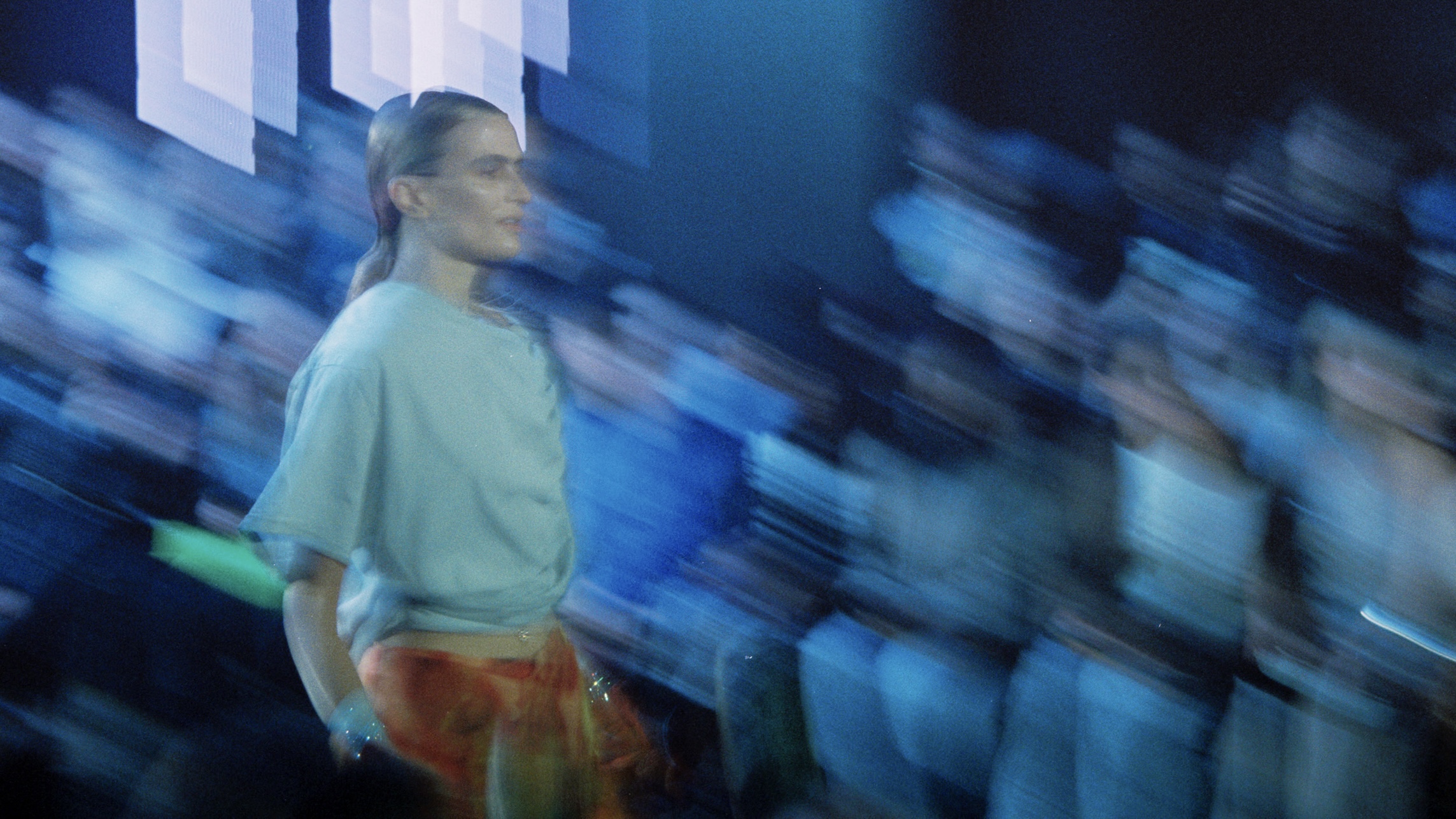 Model walking down a runway with blond hair in a ponytail, a blue t-shirt, and orange pants.