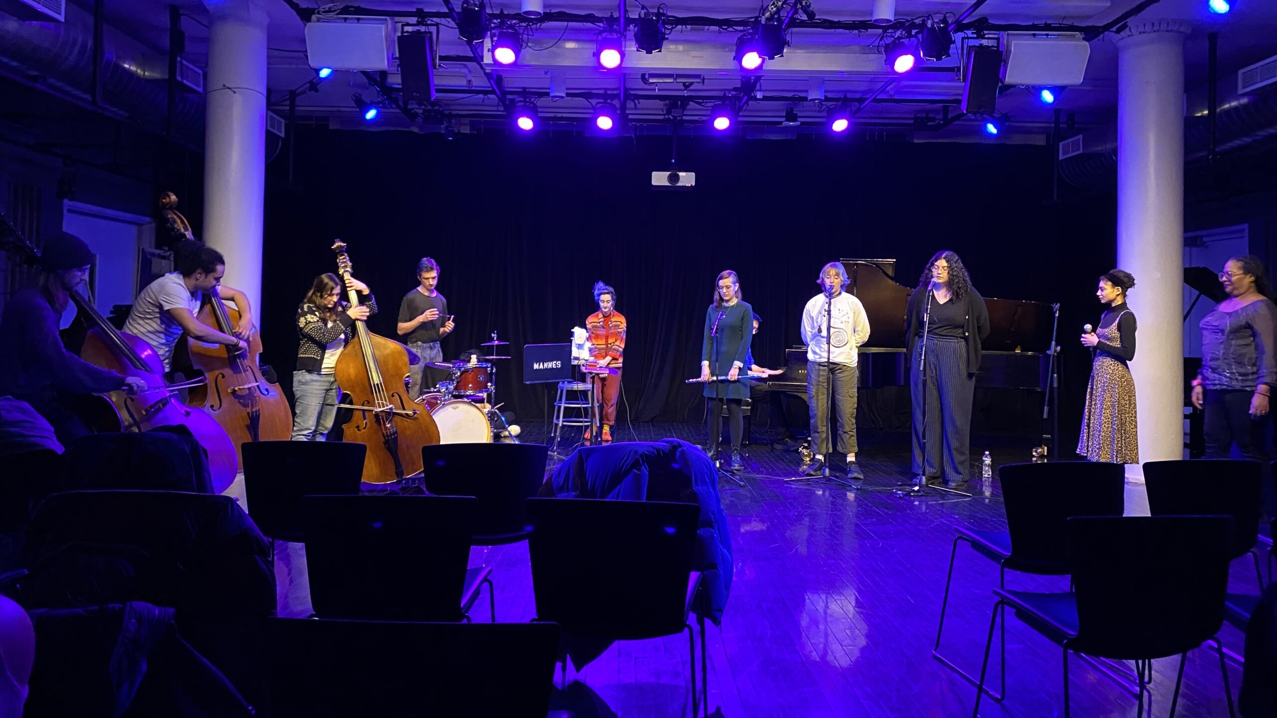 11 students and alumni stand on stage and perform with microphones, basses, and a drum set with rows of chairs in front of them.
