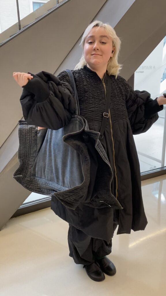 Student stands with arms bent and open at the shoulders wearing a ruched zip-up black dress with poofy sleeves, loose black pants, black shoes, and a large denim bag with pointed corners.