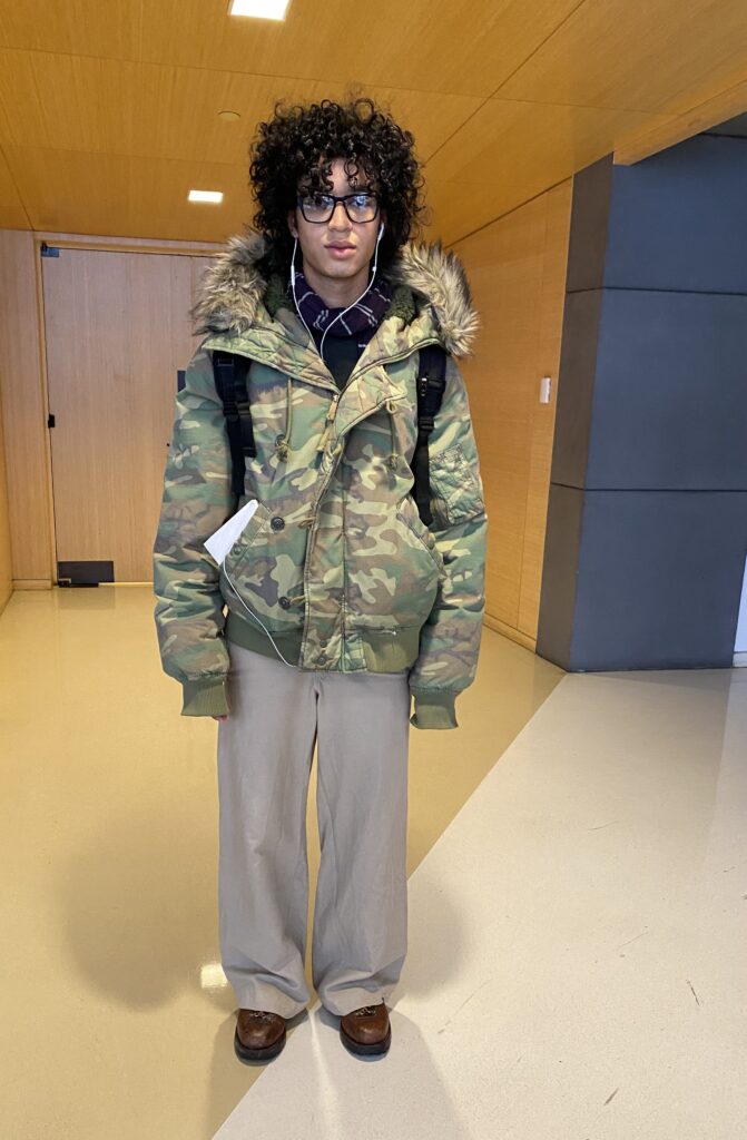 Student wears a camo puffer jacket with a fur-trimmed hood, a purple and white striped scarf, wide-leg cream colored pants, brown leather shoes, and a black backpack