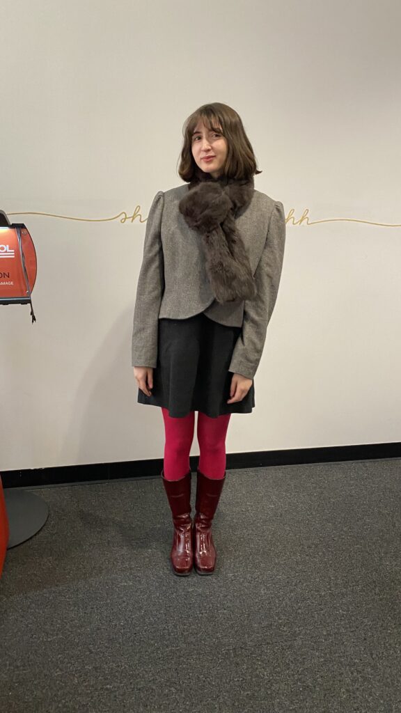 Student wears a rabbit fur scarf, cropped gray jacket, black mini skirt, red tights, and burgundy patent boots.