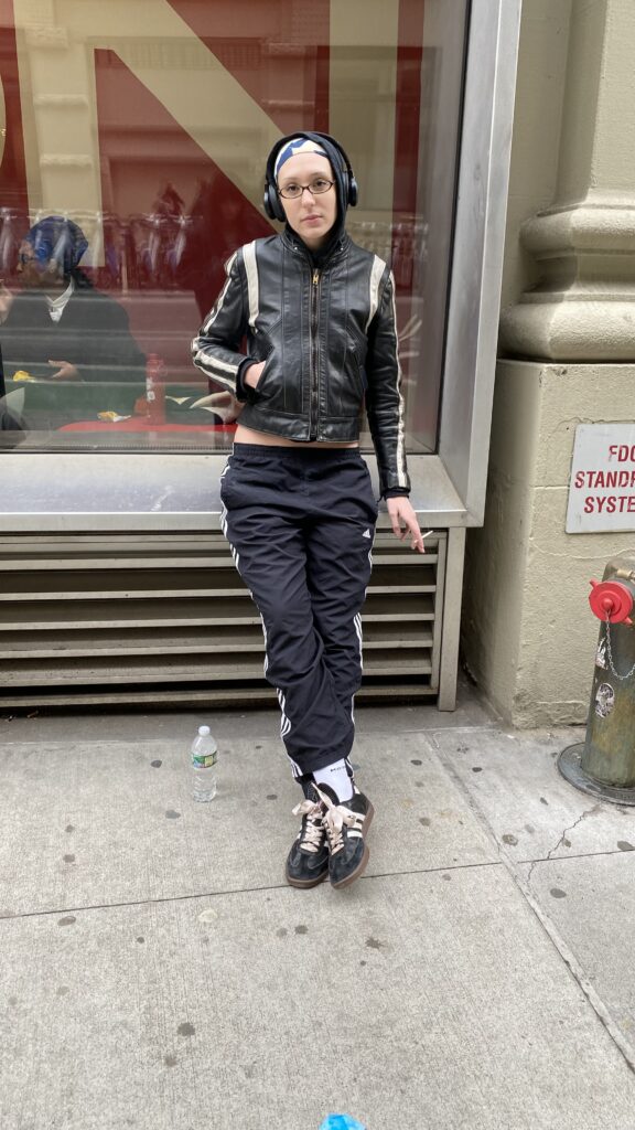 Student wears black headphones, a blue and white beanie, moto jacket, black Adidas track pants, and Adidas sambas tied with light pink ribbons.