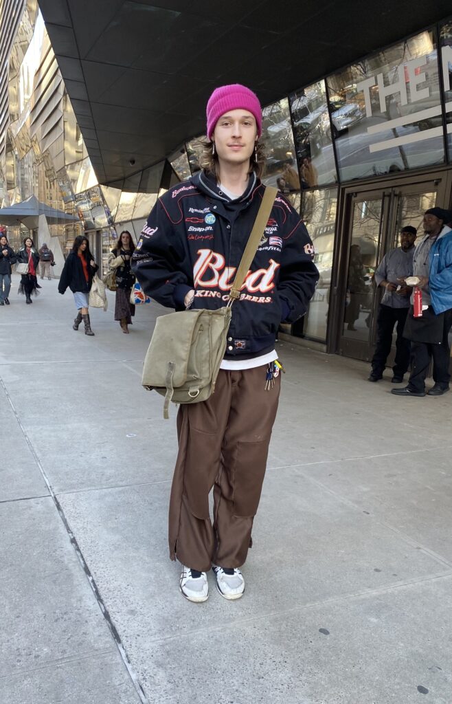 Student wears a magenta beanie, long dangly earrings, a Budweiser jacket, brown pants, white sneakers, and an army green crossbody bag.