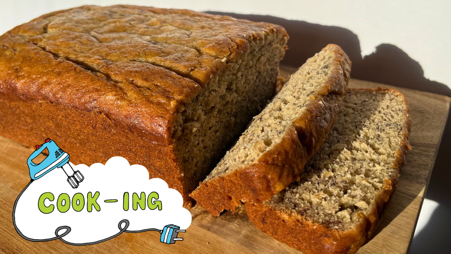 A partially sliced loaf of banana bread sitting on a brown cutting board with a white background. A logo in the bottom left corner reads “Cook-ING” with a blue hand-mixer.