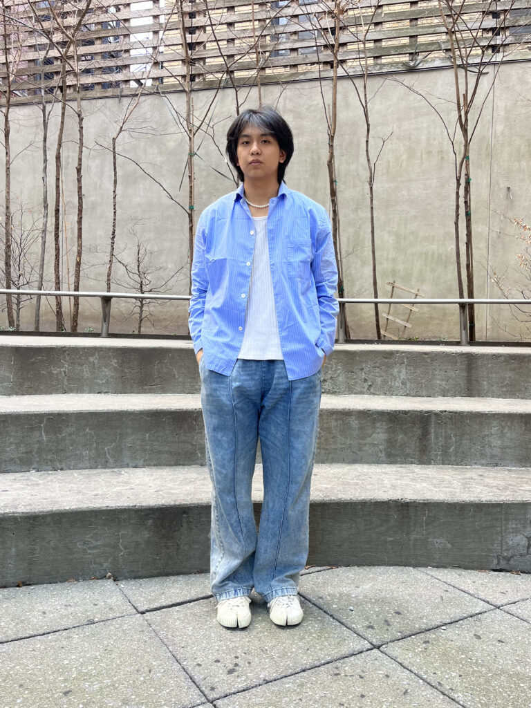 Student wears a pearl necklace, blue button up shirt open to show a white undershirt, blue jeans, and white tabi sneakers while standing in Lang courtyard.