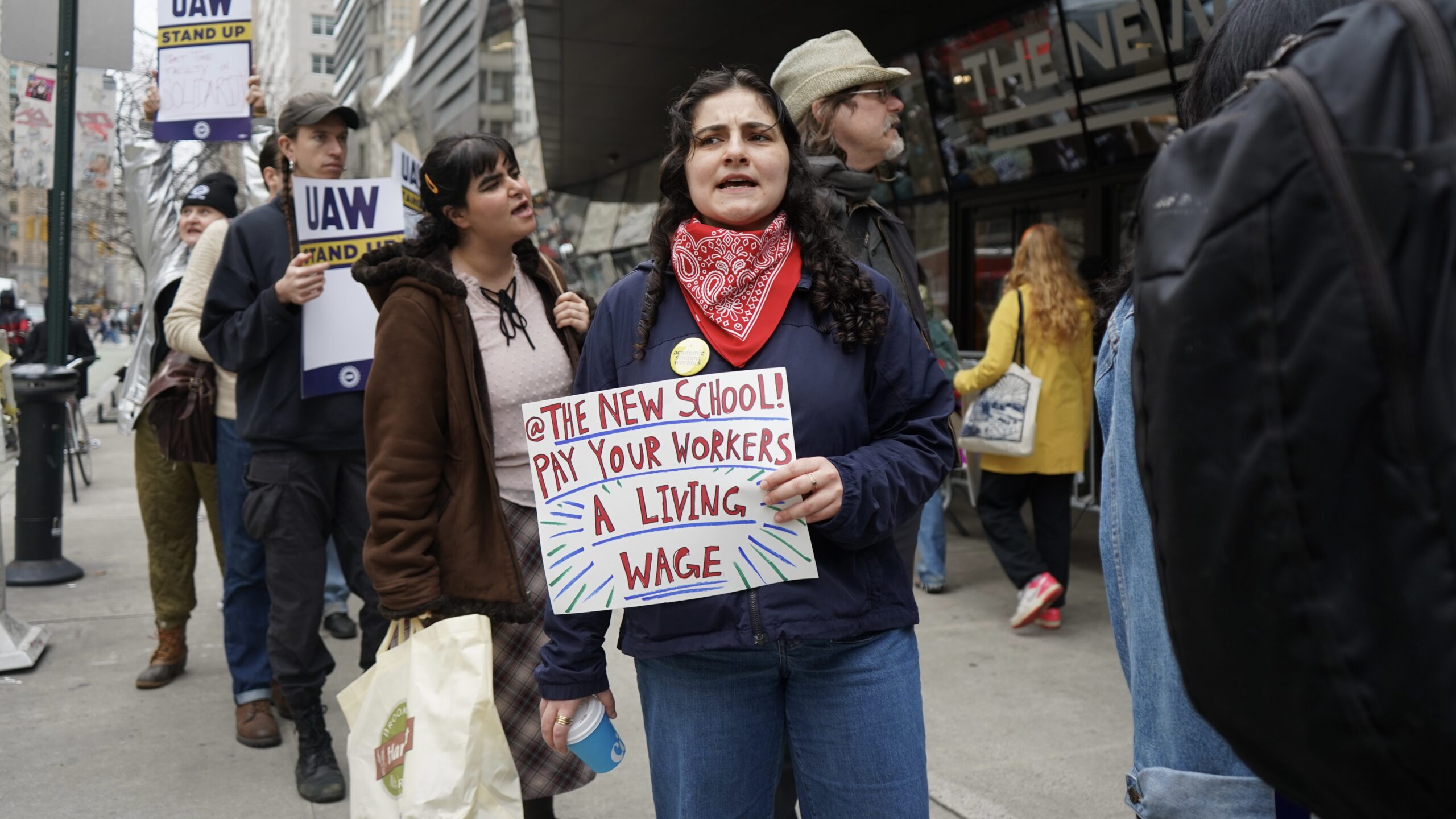 Image of a picketer outside of The New School University Center holding up a sign that reads “@ The New School! Pay your workers a living wage.” More protesters are seen in the background walking along the picket line chanting and holding UAW union posters.