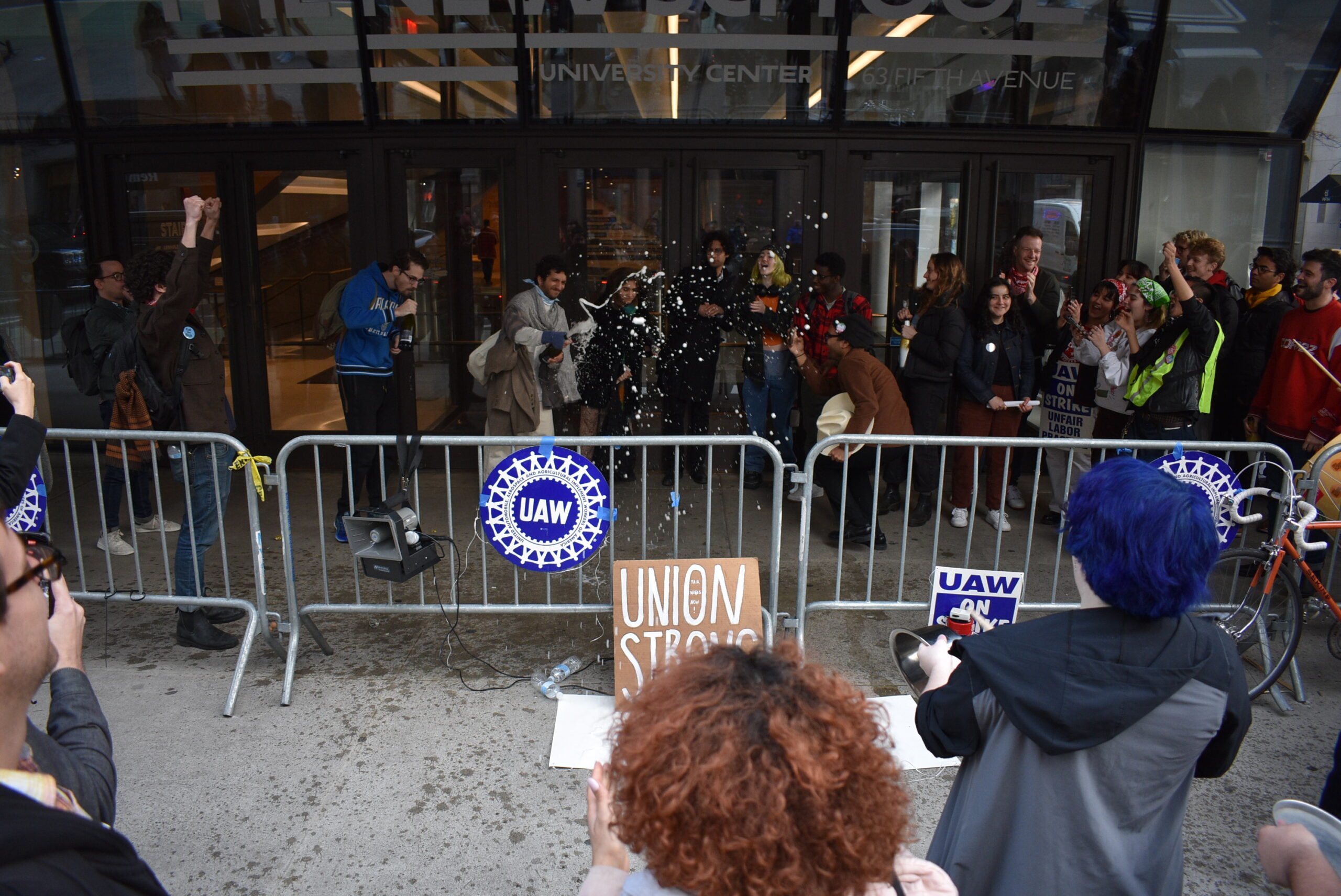 Union members celebrate with champagne in front of the UC, where temporary metal fences mark the UAW picket line.