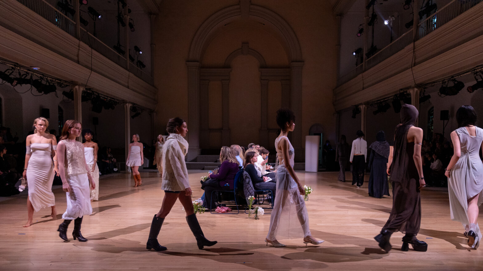 Models dressed in various designs walk under the spotlights from left to right in a circle inside St. Mark’s Church. In the center of all the models are people sitting in chairs back-to-back, forming a line.