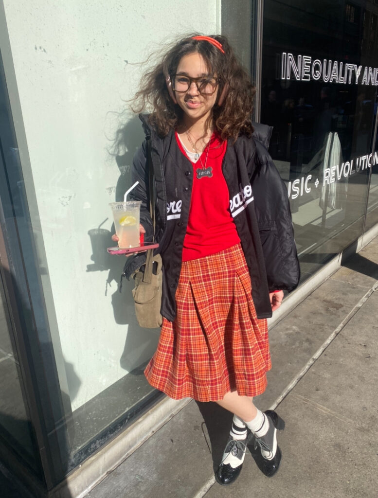 Student stands in front of the University Center wearing brown glasses, a red sweater, a red and orange plaid skirt, a black puffer coat, a red headband, and black and white saddle shoes.