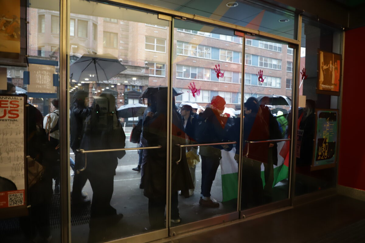 The view from inside Arnhold Hall shows protesters unfurling a Palestinian flag. On the glass doors at the entrance of the building three handprints left in red paint are visible.