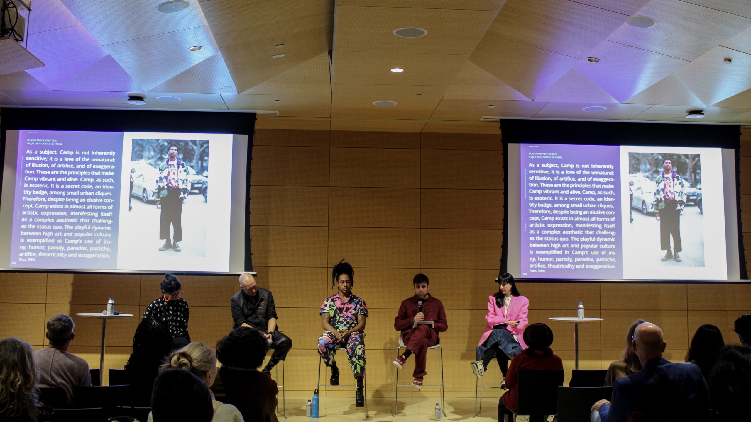 (from left to right) Lucia Cuba, Otto von Busch, MX Oops, Dirk Reynders and Marie Genevieve Cyr sit side-by-side on stage in the Starr Foundation Hall as they speak to an audience. Behind them are two large screens with an excerpt from “Fashion+.”