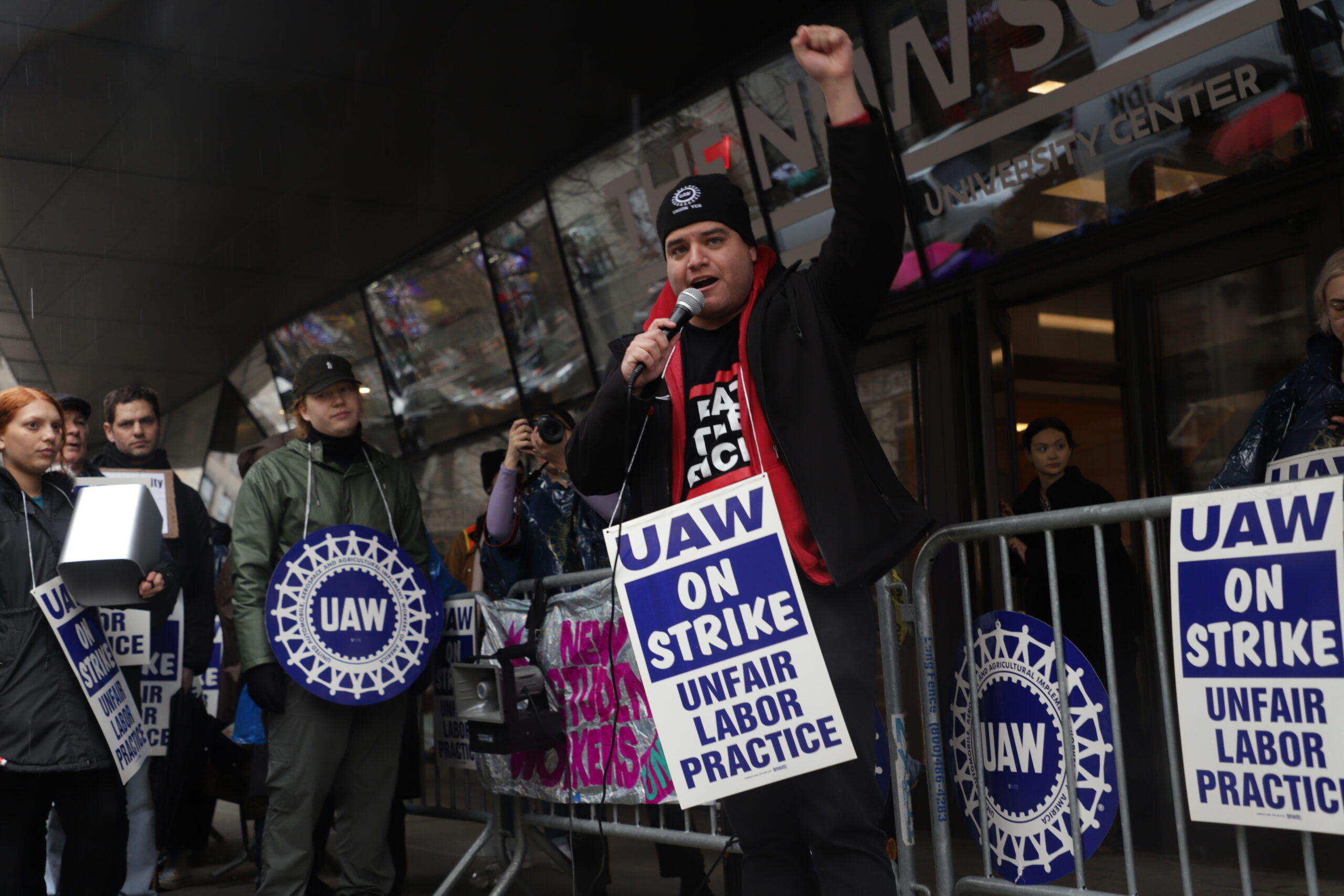 Image of a man holding his fist up as he speaks into a microphone in front of the University Center. Around his neck hangs a sign. It reads “UAW on strike. Unfair labor practice.”