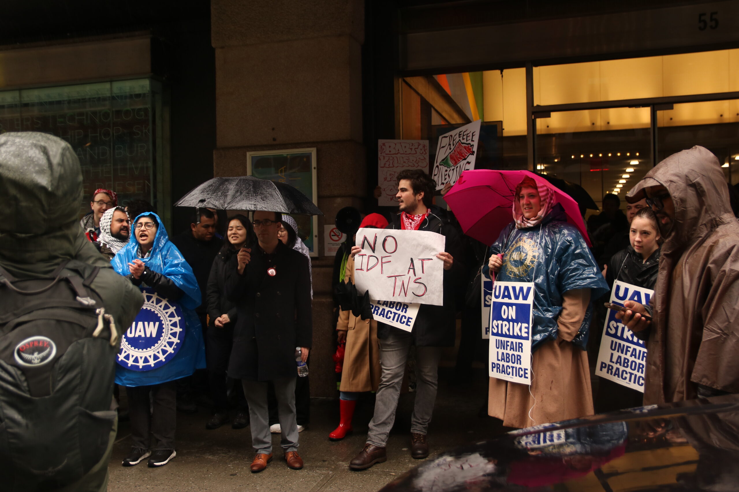 Protestors and picketers stand outside the main entrance to Arnhold Hall. Some are wearing ponchos and raincoats, and others holding umbrellas. A few people are wearing signs that say “UAW On Strike, Unfair Labor Practice.” An individual in the center of the image, wearing a red bandana around their neck, is holding a sign that reads, “NO IDF AT TNS.”
