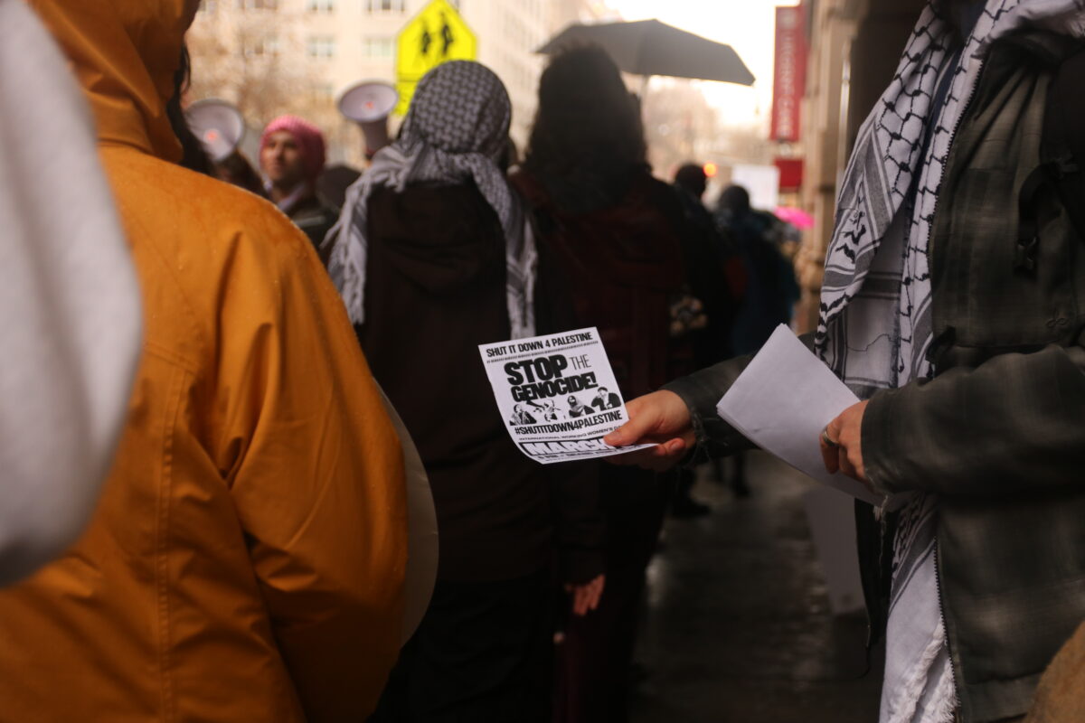 On the right side of the image, the torso of a figure wearing a flannel and black and white keffiyeh is visible. They are handing out flyers that “Shut It Down 4 Palestine, Stop the Genocide!” to an individual on the left side of the image, wearing a yellow jacket with the hood up. 