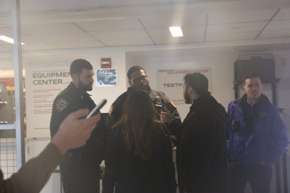 A slightly foggy photo shows two uniformed NYPD officers and one Community Affairs officer speaking to students who have their backs to the camera. In the foreground is the forearm of a person on their phone.