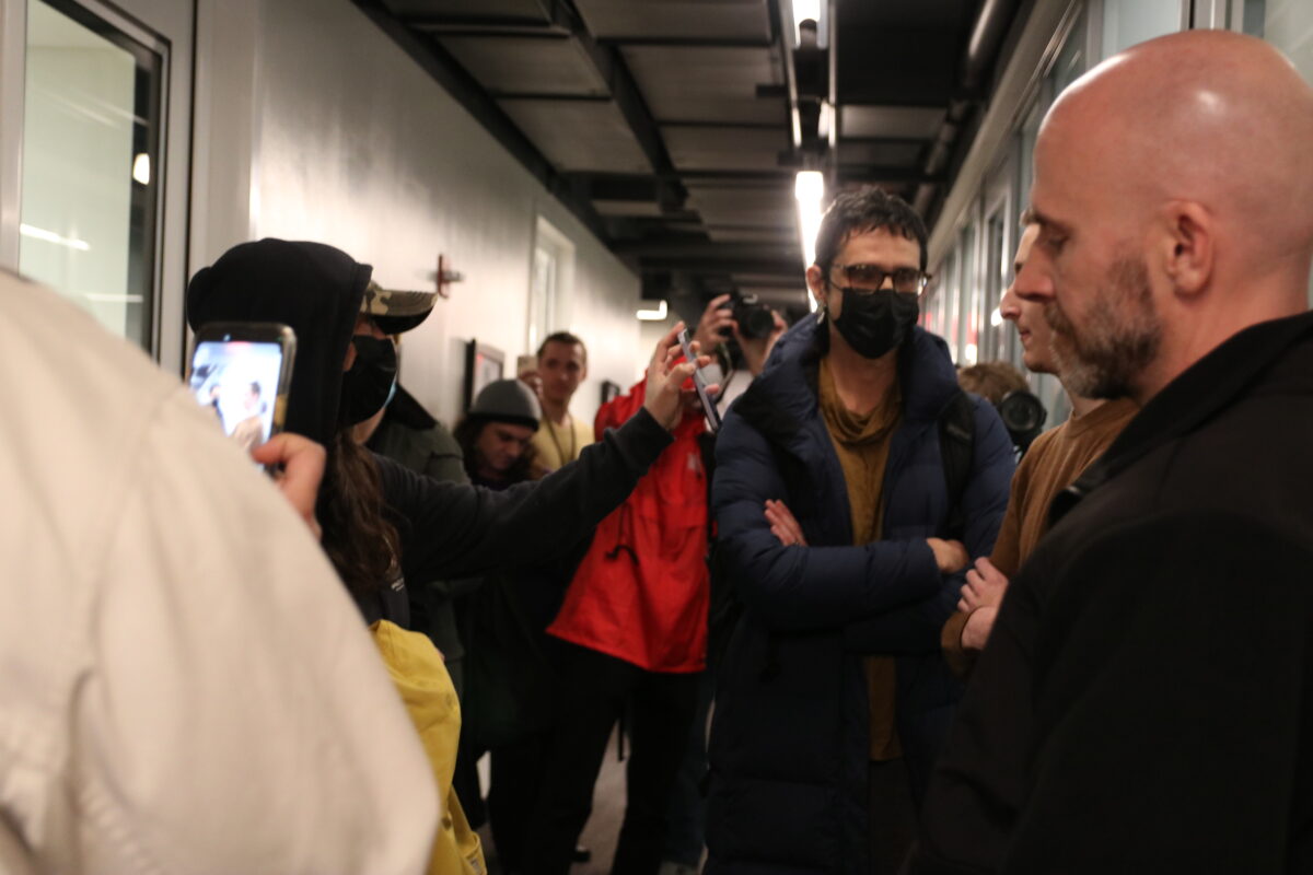 A protestor wearing a baseball cap, black hoodie, and black face mask showing her phone to an individual standing at the door to Richard Kessler’s office. In the background, another protestor stands with their hands crossed. An individual with a red jacket has their arms raised taking a photo of the interaction.