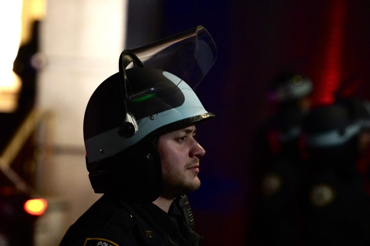 A male NYPD officer wearing a riot helmet looks stoically towards the crowd 