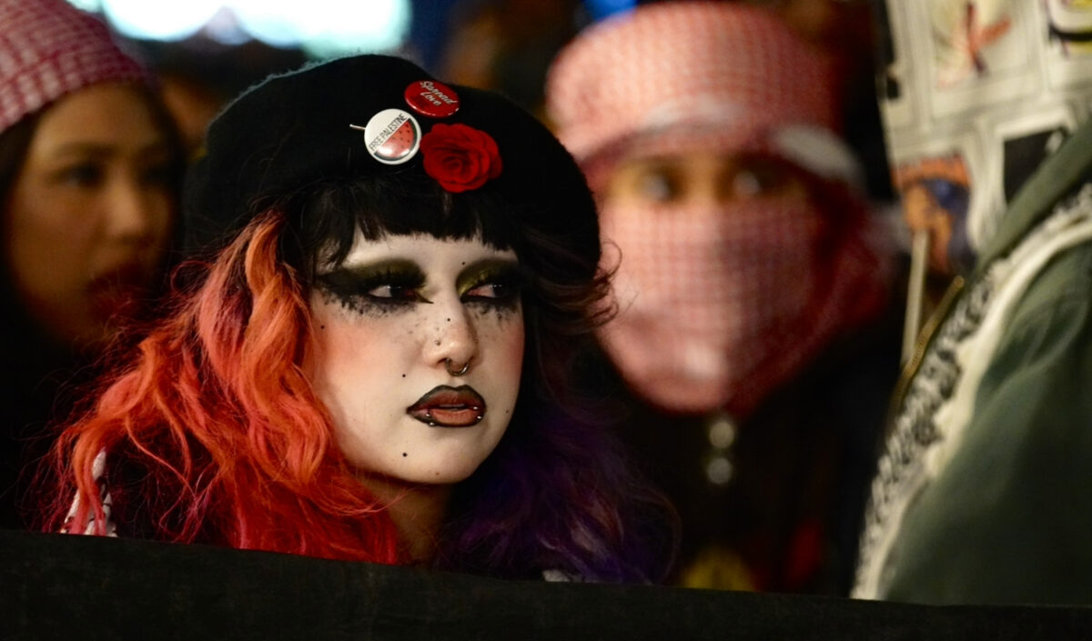 A girl with half/half red and purple hair with her face painted white wearing a black hat with a red flower and two pins