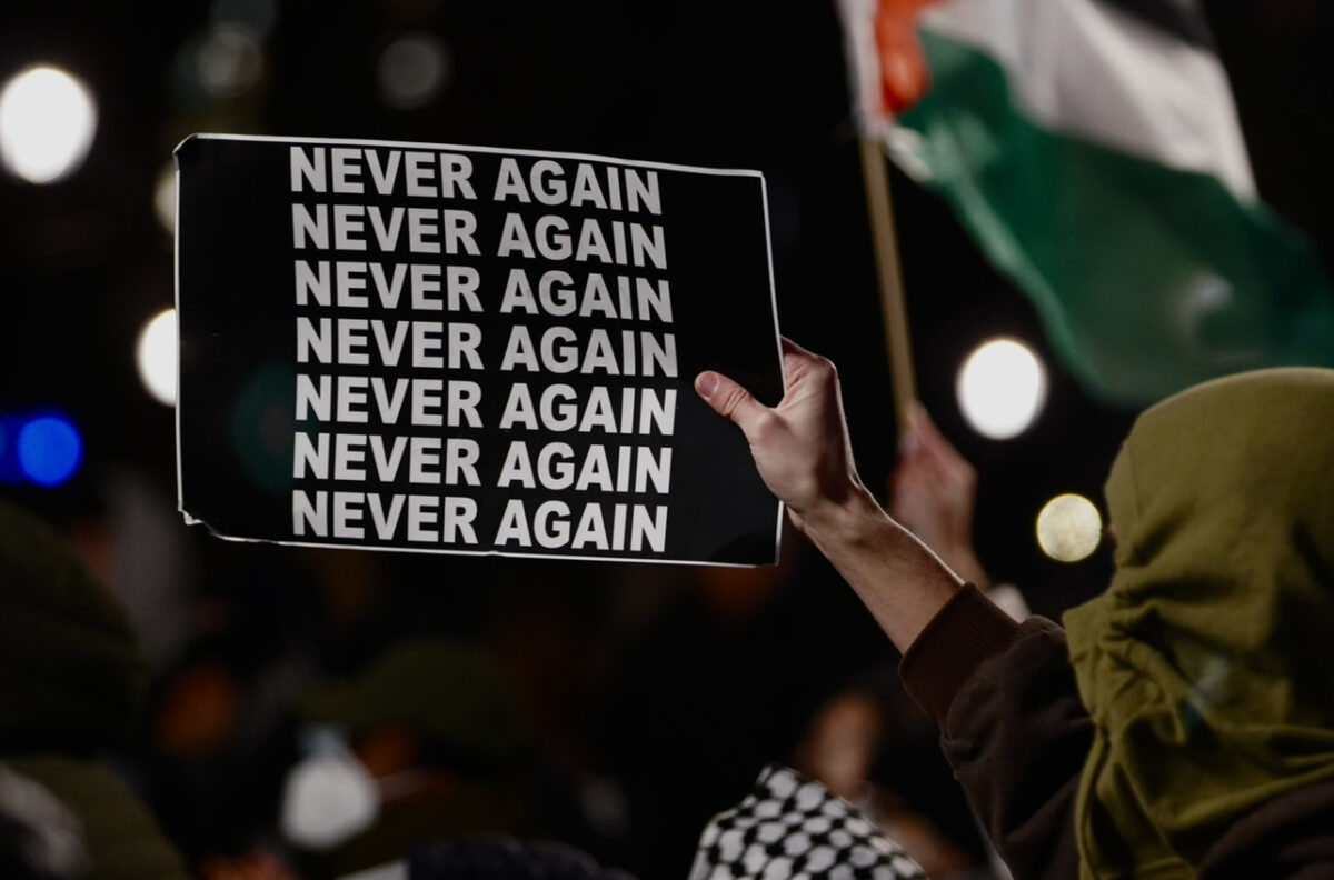 A protester with their face concealed by an olive mask holds a black sign with the words “Never Again” repeated seven times 