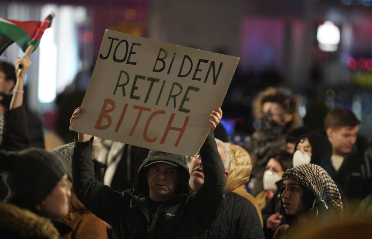 A man dressed in all black holds a sign that reads “Joe Biden Retire Bitch” in black, green, and orange letters