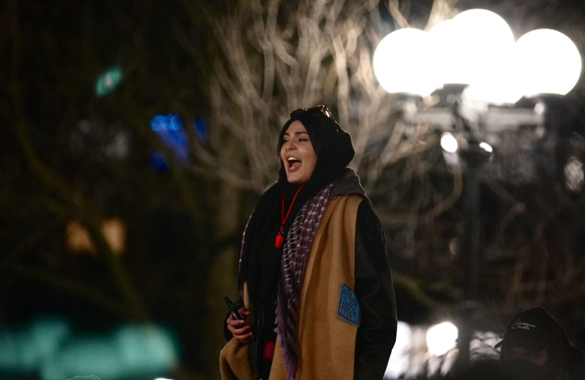 A woman wearing a black hoodie, a dark purple keffiyeh, a kahaki scarf, and red whistle around her neck yells out toward the crowd 