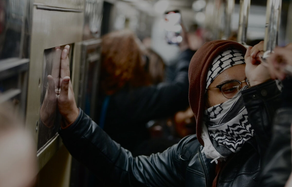 Protester wearing a black leather jacket, a brown hoodie, glasses and a, keffiyeh hitting his hand against the wall in a train packed with protesters