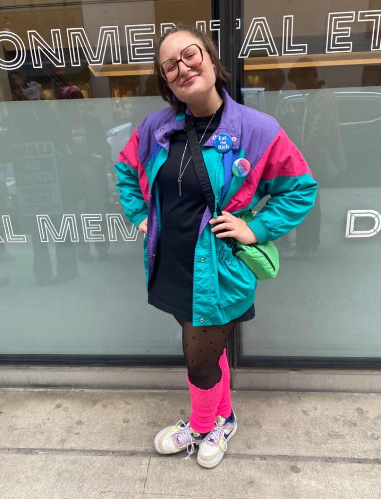 Student stands in front of the University Center wearing glasses, a purple, hot pink and teal colored windbreaker, a black mock-neck dress, black polka dot tights, hot pink leg warmers, white and lavender Nike Air Force Ones, and a green fanny pack.