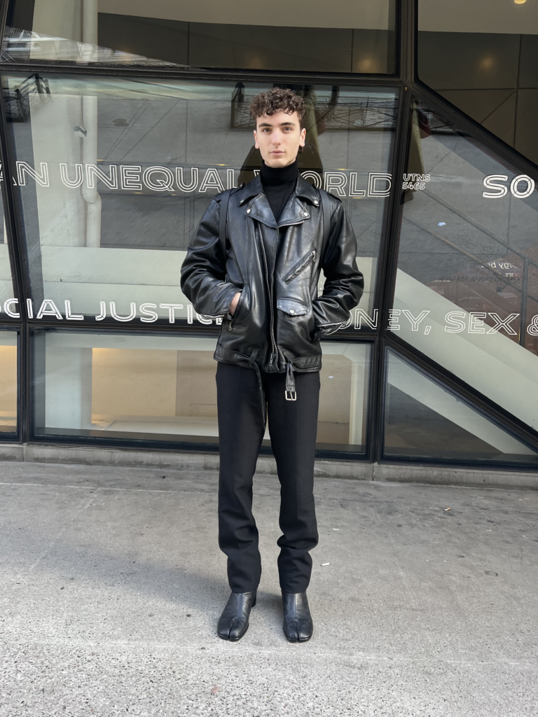 Student wears a black turtleneck, black trousers, a black leather jacket, and black leather tabi boots while standing in front of the University Center.