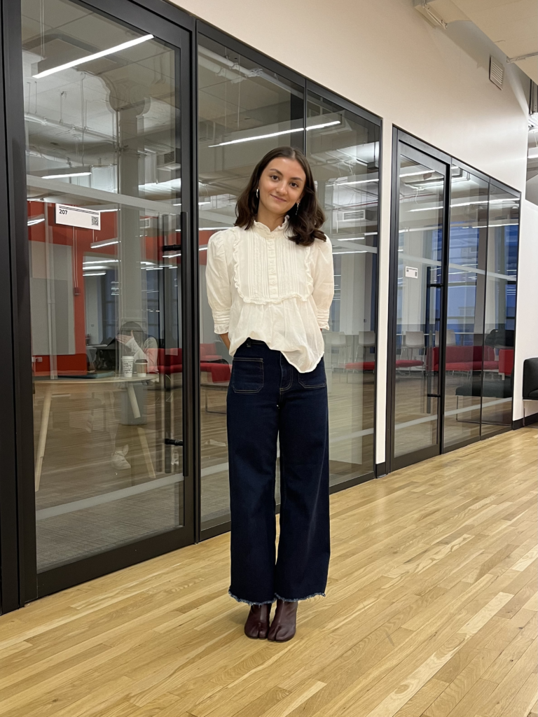 Student stands in the 13th St. building wearing a white frilly top, dark wash denim jeans, and merlot colored tabi boots.