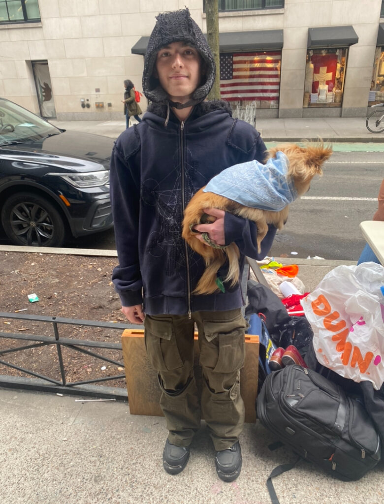 Student stands on the sidewalk across from the Brandy Melville on 14th Street wearing a navy blue hat secured by a strap, a navy blue zip-up hoodie, green cargo pants, and black sneakers, while holding a Pomeranian that’s wearing a baby blue Student Worker Power bandana.