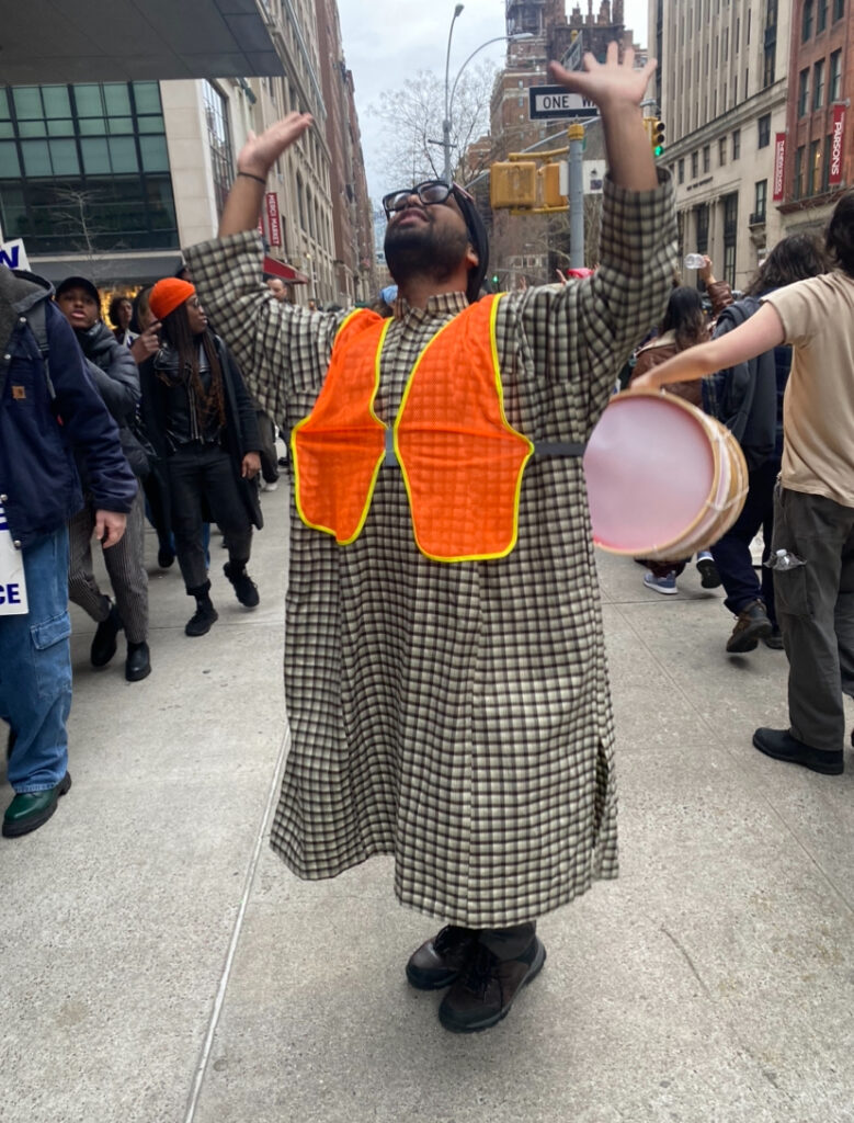 Student stands in the middle of the picket in front of the University Center wearing a black and gray pheran, a mesh fluorescent orange construction vest, a black beanie gray pants, and dark brown hiking boots.