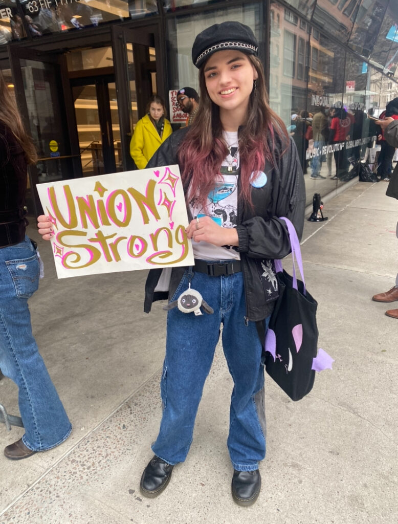 Student stands in front of the University Center wearing a black beret with a chain-link, a black leather jacket, a white graphic T-shirt, dark wash jeans with a lamb keychain clipped to one of the belt loops, a pair of black Doc Martens, a purple and black tote bag holding a sign that reads “Union Strong” in gold and pink lettering.