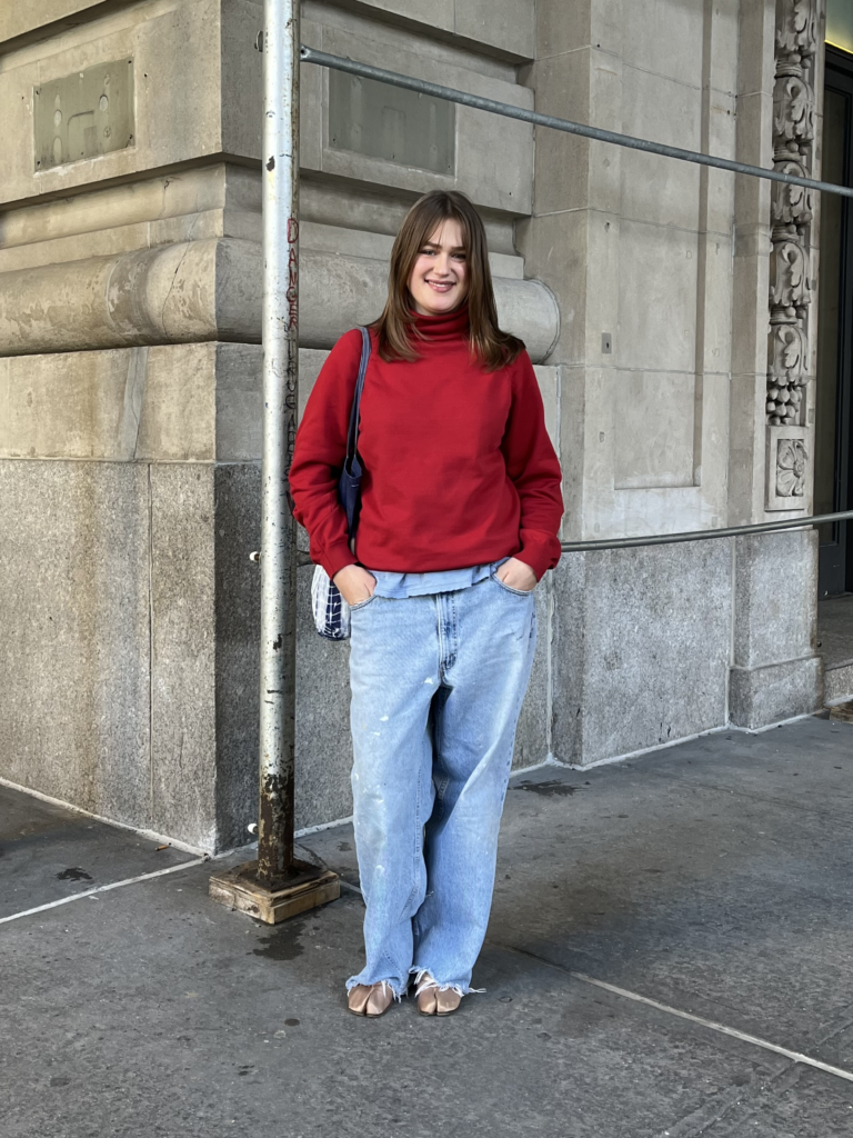 Student wears a red turtleneck sweater, blue jeans, a tie-dyed blue bag, and pink tabi satin ballet flats while standing on the corner of 16th St and Fifth Avenue.