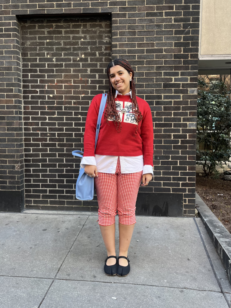 Student wears a red sweater layered over a white button up shirt, red gingham capri pants, black Mary Jane tabi shoes, and a baby blue Telfar tote bag while standing in front of a black brick wall.