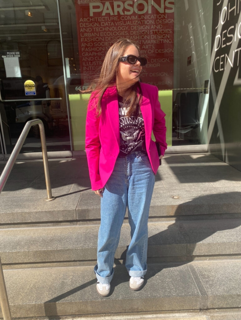 Student stands on the steps in front of the Parsons 13th Street building wearing a hot pink blazer, a black graphic T-shirt, medium wash baggy jeans, and a black and white pair of Adidas sneakers.