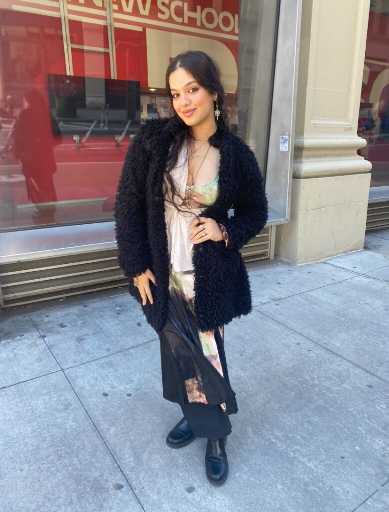 Student stands in front of the Parsons 13th Street entrance wearing an abstract green, light brown, and black tie-dye dress, black boots, a black faux fur coat, and gold star earrings.