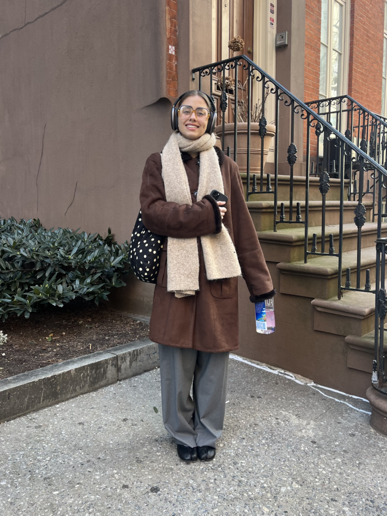 Student stands in front of a brownstone wearing round glasses, gray headphones, an oatmeal knit scarf, brown coat, gray trousers, black leather ballet flats, and a black-and-white polka dot bag.