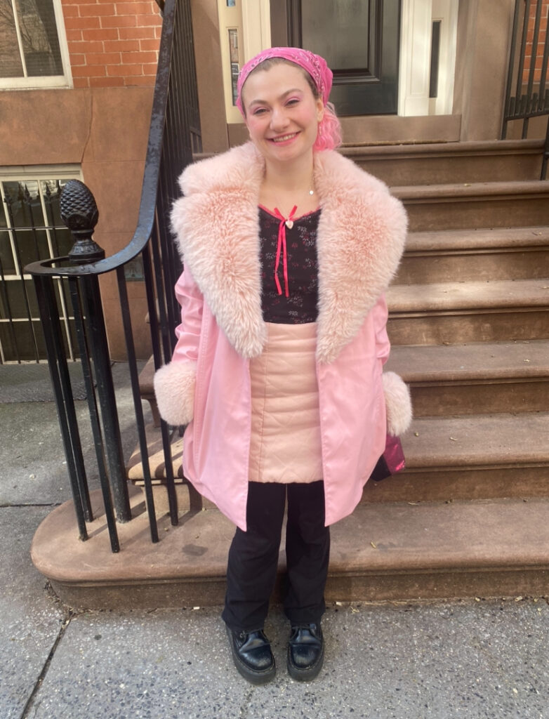 Student stands in front of the steps next to the Lang 12th Street building wearing a pink coat with a faux fur collar and cuffs, a black top with a hot pink bow, a light pink quilted skirt, black pants, a black pair of Doc Martens, and a pink bandanna to tie their hair back.