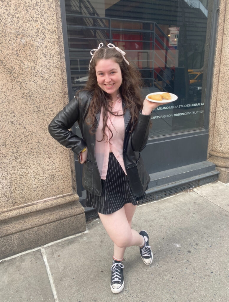 Student stands in front of The New School Welcome Center on 5th Avenue holding a plate of empanadas wearing a black leather blazer, a pink button-up shirt, black and white pinstripe shorts, and a pair of black and white high-top Converse.