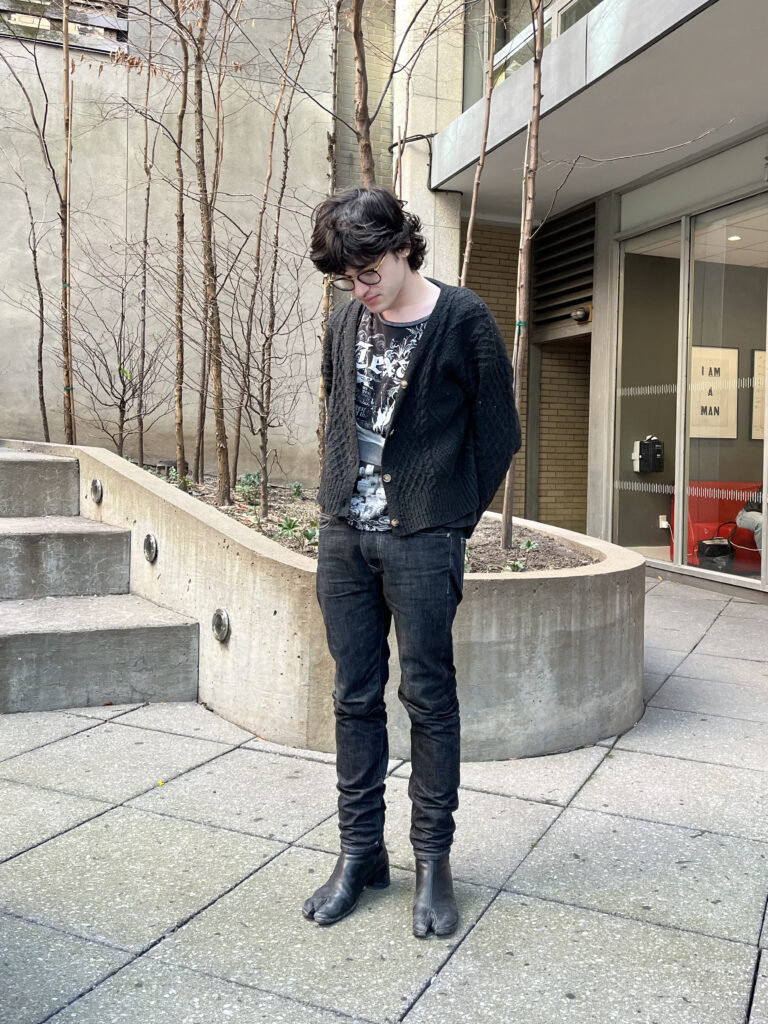 Student stands in the Lang courtyard wearing a black cardigan, graphic shirt, black skinny jeans, and black heels tabi boots.