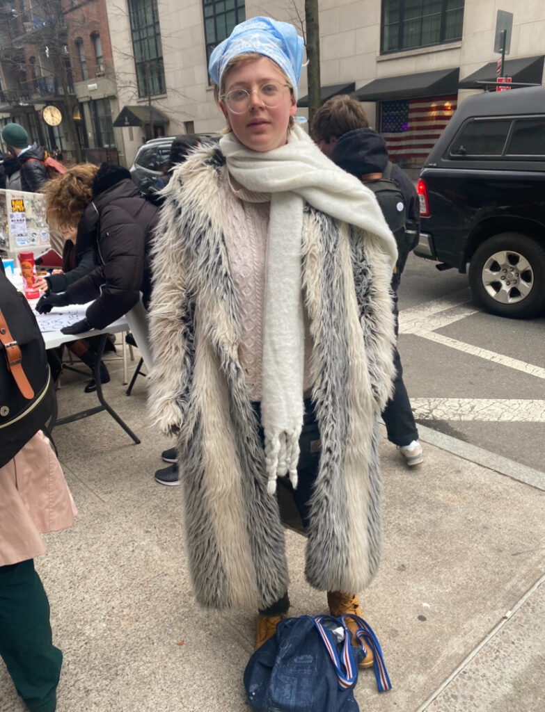 Student stands on the sidewalk across from the Brandy Melville on 14th Street wearing a baby blue Student Worker Power bandana, a chunky white scarf, a longline gray and cream-colored faux fur coat, a light pink sweater, dark wash jeans, and lace-up camel boots.