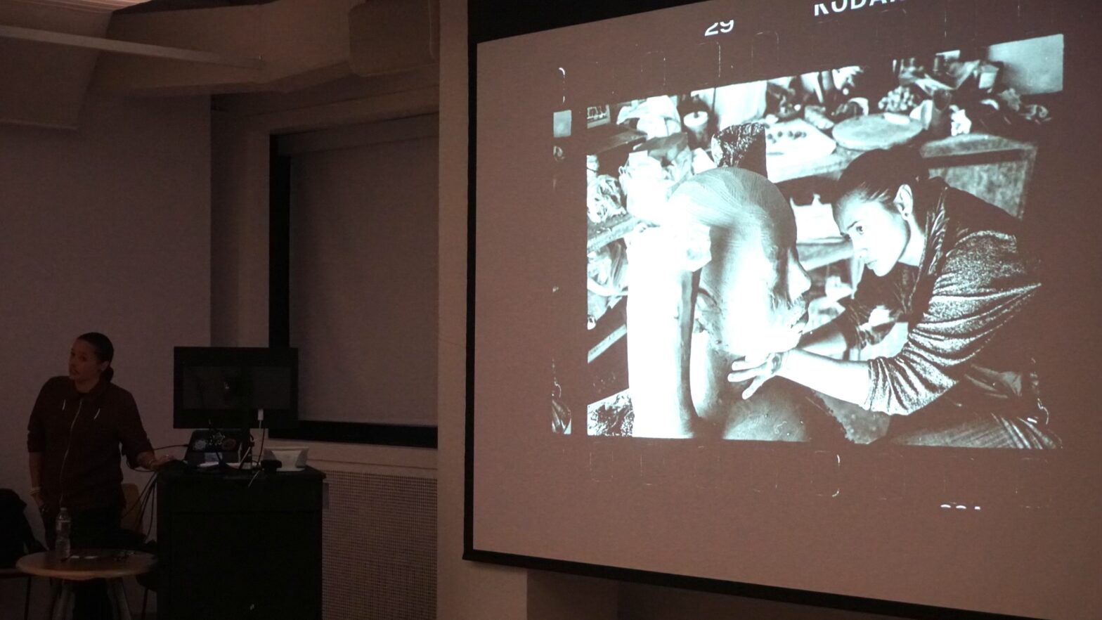 In a dimly lit lecture room, Rose B. Simpson stands in front of a projector that portrays a picture of her carving one of her signature clay statues.