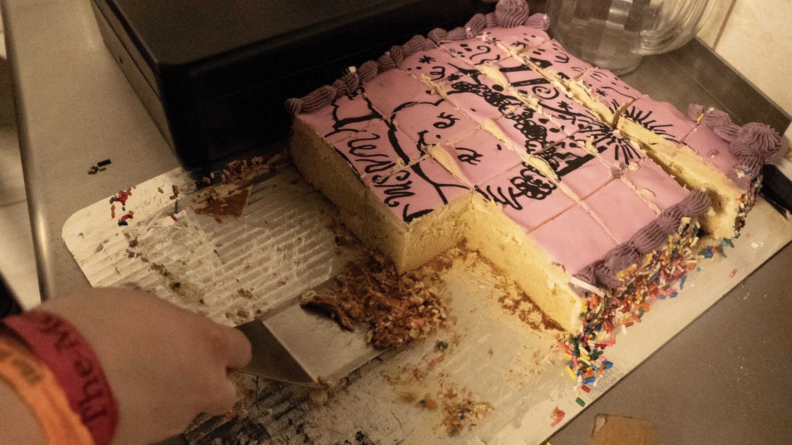 Image of a half eaten cut up birthday cake. The cake is pink with an angel wearing a WNSM shirt drawn in icing on top
