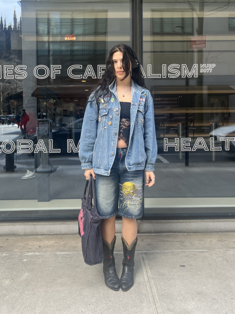 Student stands in front of The New School University Center wearing a denim jacket, navy lace crop top, embroidered jean shorts, black cowboy boots, and a black denim bag.