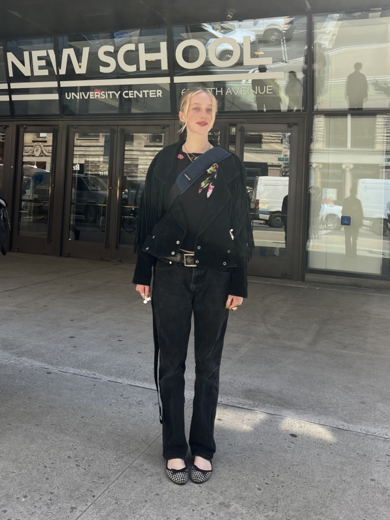 Student stands in front The New School University Center wearing a black suede fringe jacket, black shirt, black jeans, and black gingham ballet flats.